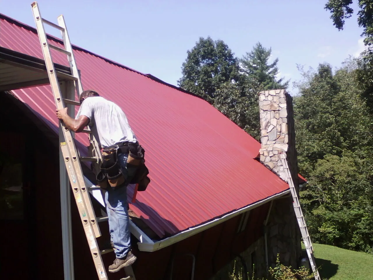 Roofer on a ladder beside a red metal roof and stone chimney — Soffit & Fascia Repair by Roofing Brothers in Brimfield